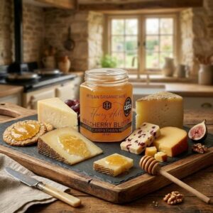 A jar of Honey Hills Wild Cherry Blossom Artisan Organic Honey from the Carpathian Mountains (227g) with a Great Taste award badge, displayed on a slate and wood cheeseboard with various cheeses drizzled in honey, a cranberry cheese, oat crackers, a honey dipper, fresh figs, walnuts, and red grapes, set in a stone-walled farmhouse kitchen.