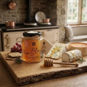 A jar of Honey Hills Plum Blossom Premium Honey (227g) with a raffia bow and Great Taste award badge, displayed on a rustic live-edge wooden board with a honey dipper, blue cheese drizzled in honey, a log of ash-rinded goat's cheese, seeded crackers, red grapes, rosemary sprigs and a wooden bowl of crackers, with an Aga range cooker and copper pans in the background.